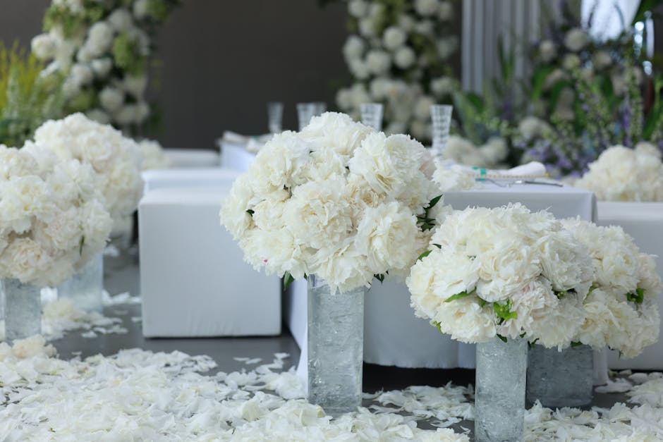 A floral arrangement featuring large, lush bouquets of white hydrangeas and roses placed in tall, cylindrical glass vases with a textured surface. The bouquets are densely packed, showcasing their sof