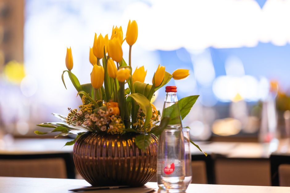 A floral arrangement featuring bright yellow tulips with smooth, elongated petals standing upright in a round, dark brown ribbed vase. Surrounding the tulips are small clusters of pink and white fille