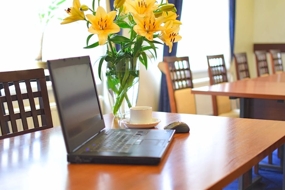 A clear glass vase containing a bouquet of vibrant yellow lilies with orange stamens and green leaves, positioned on a polished wooden conference table. The arrangement is lush and fresh, with petals 